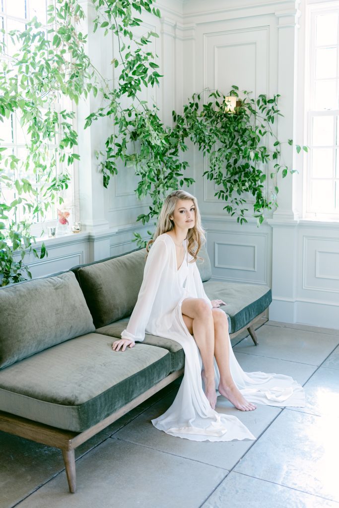 blonde model in white robe and nightgown sits on green couch surrounded by greenery