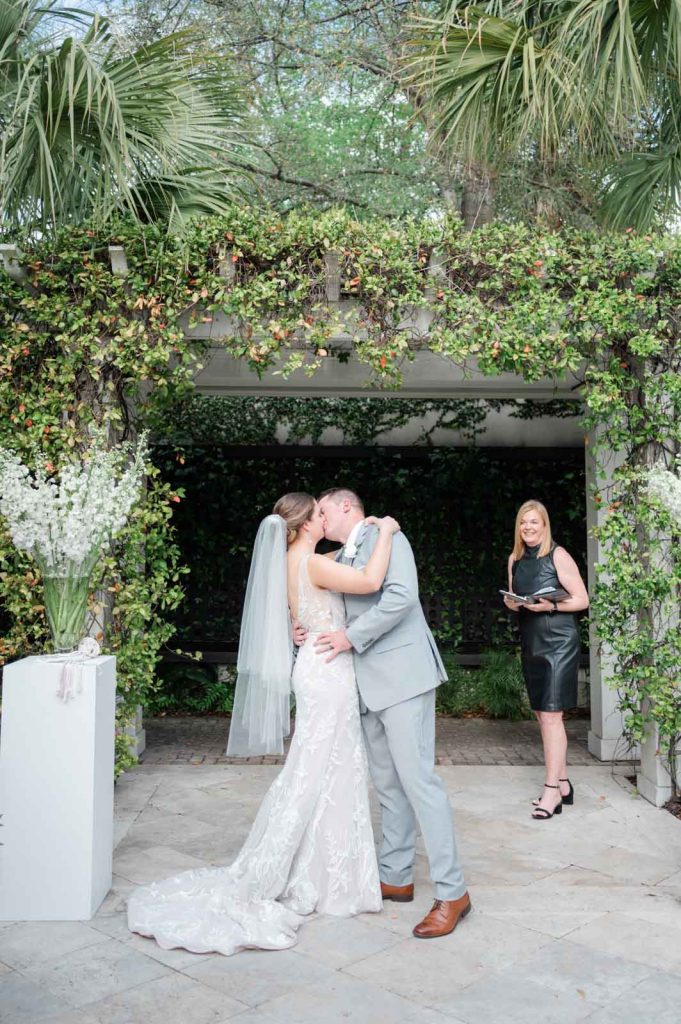 newlyweds kiss in front of arbor covered in greenery after wedding ceremony with officiant looking on