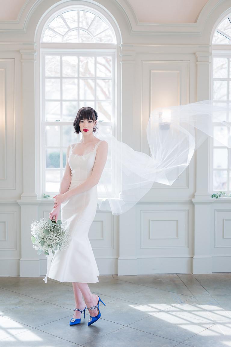 Bridal portrait of bride in tea length dress, blue shoes, and veil floating in the air