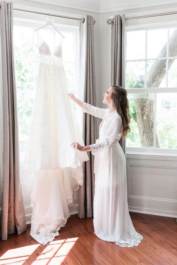 bride in white robe holding wedding dress hanging from window