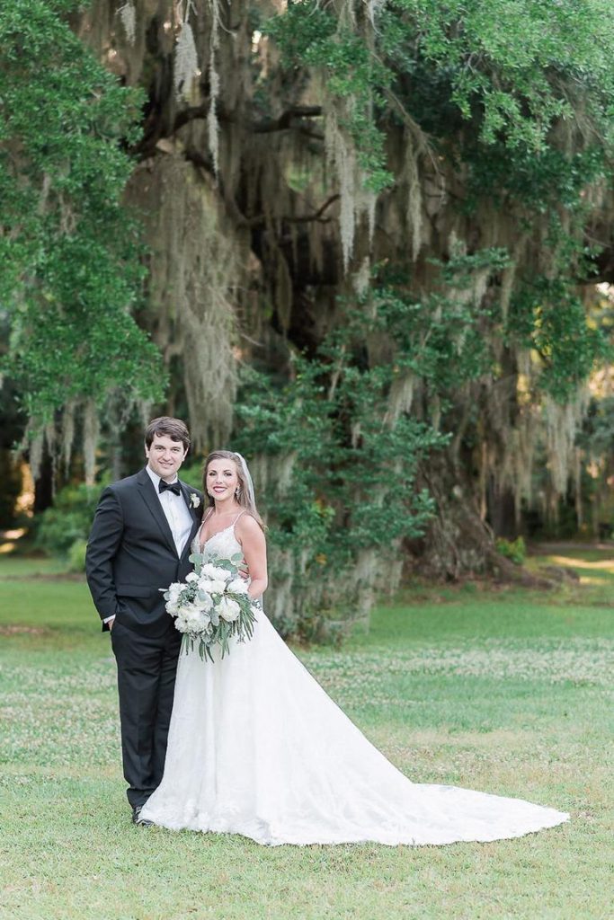 Bride and groom stand on lawn in front of oak trees with Spanish Moss