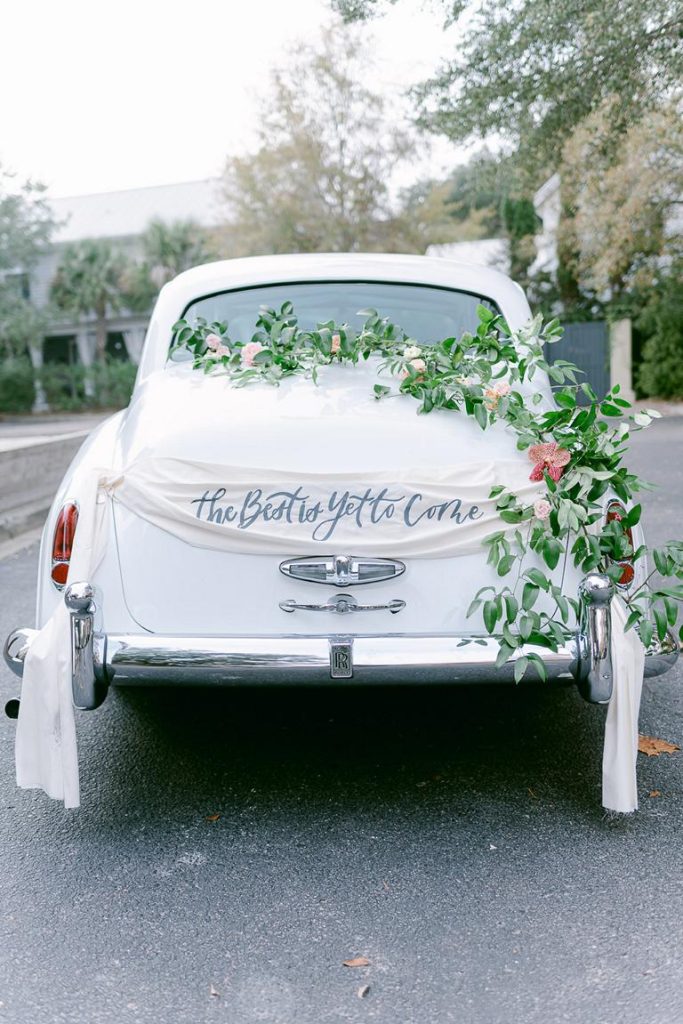 rear of White vintage Rolls Royce decorated with greenery, flowers and a banner reading " The best is yet to come"