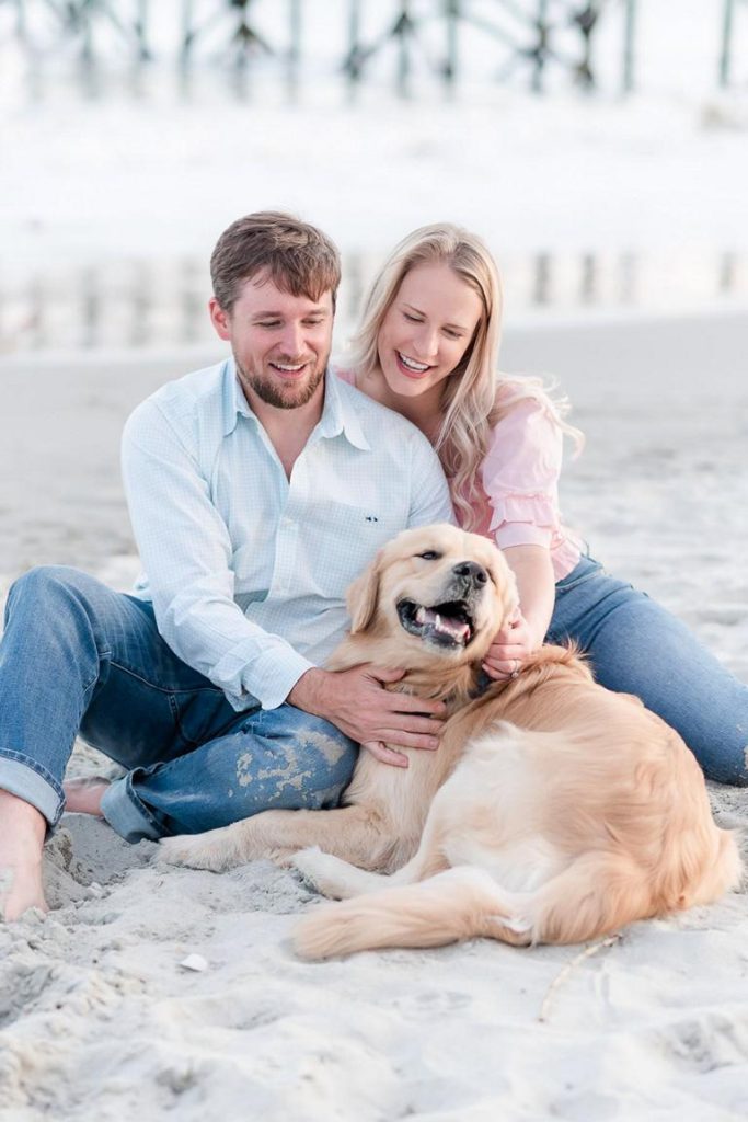 Engaged couple petting their dog while sitting at the beach