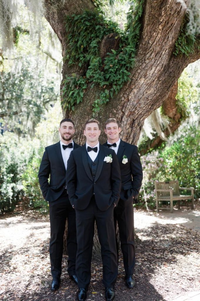 Groom with his brothers all dressed in black tuxedos standing in front of a tree with hands in their pockets