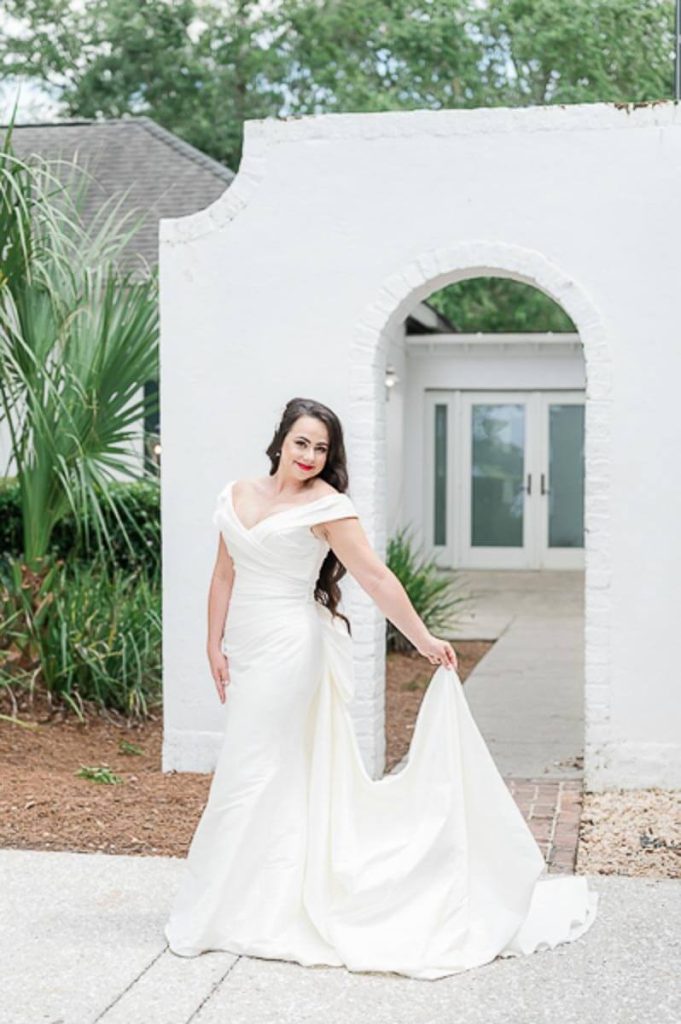 Bride holding train of dress while standing in front of curved arch entryway