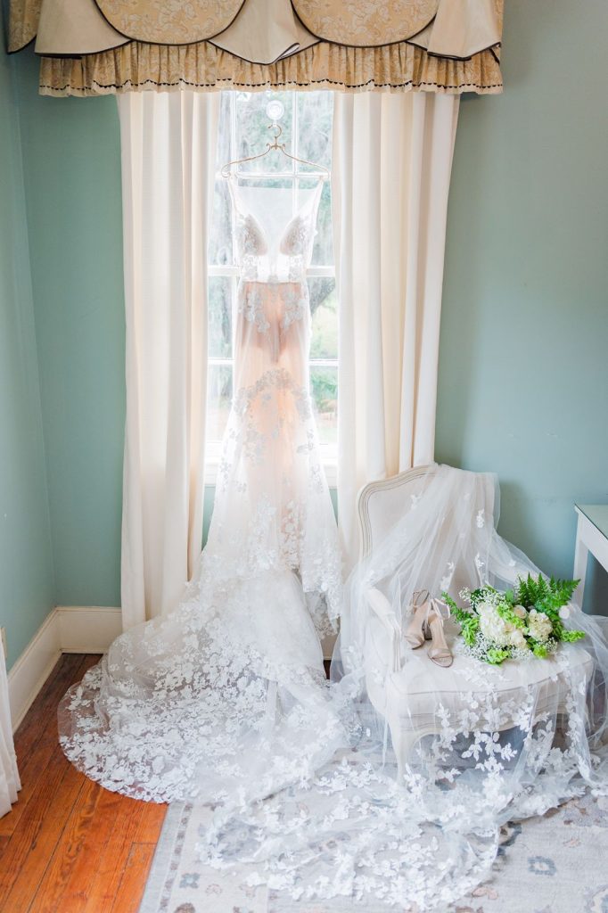 wedding dress hanging from window with veil and shoes bouquet sitting on chair nearby
