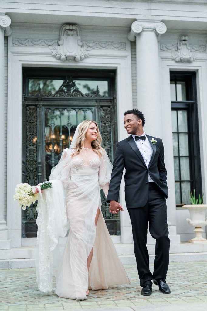 Newlyweds walk hand in hand outside of mansion