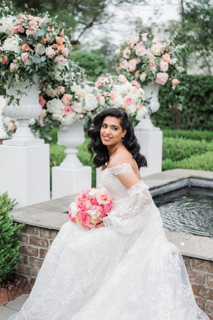 bride sitting besides fountain in garden surrounded by flowers