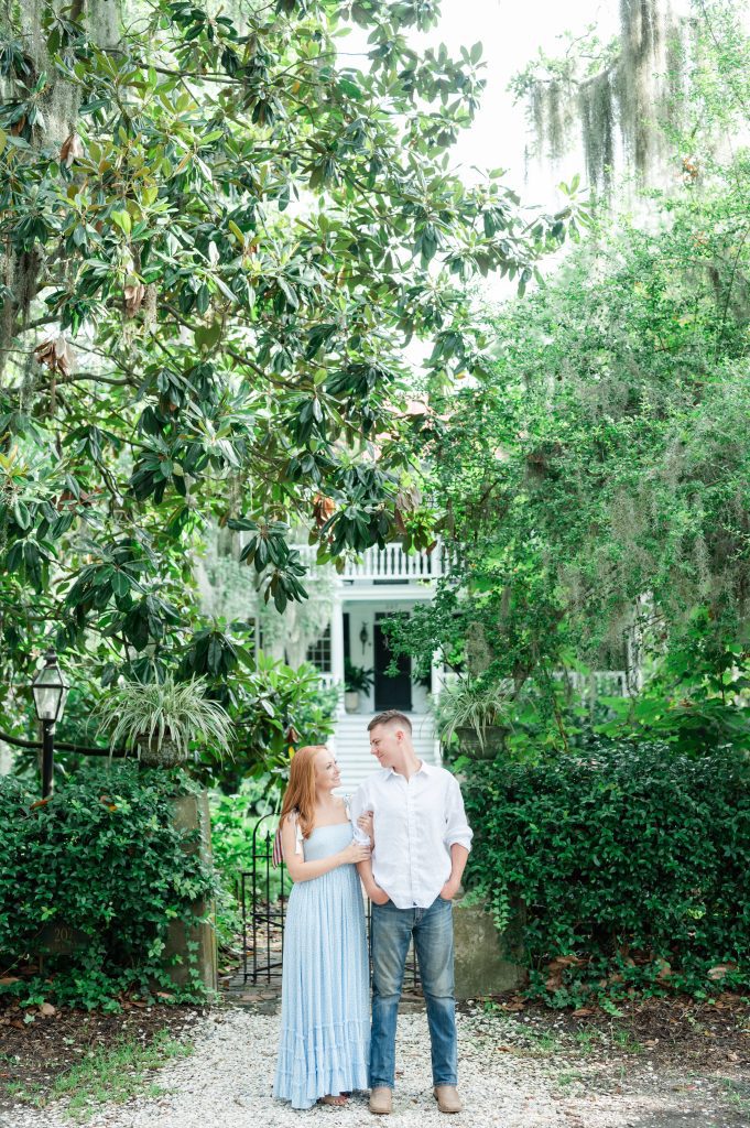 newly engaged couple stand arm in arm in front of white house & magnolia tree