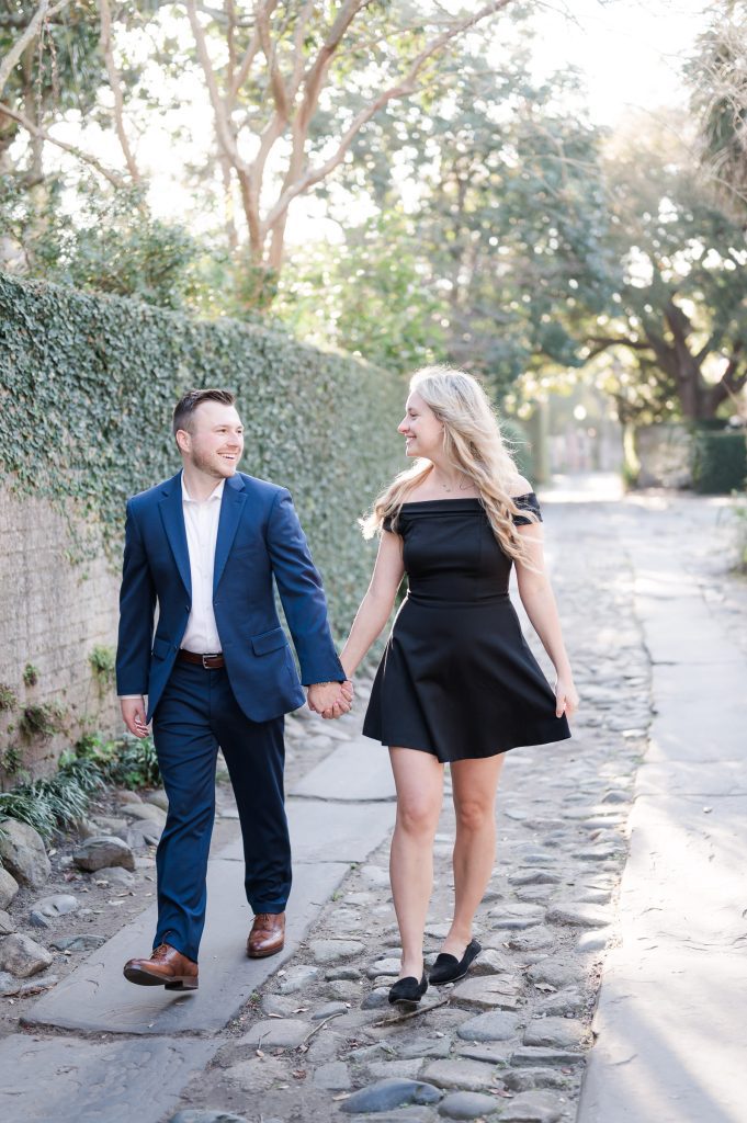 Newly engaged coupe walk hand in hand down cobblestone alley in Charleston, SC