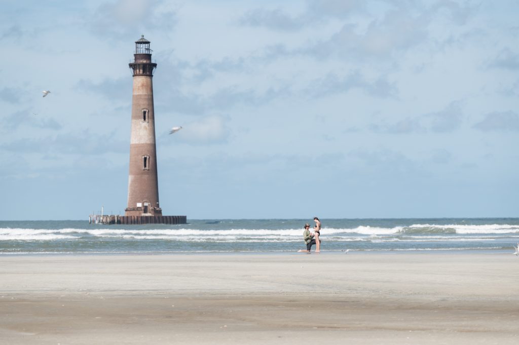 guy on knee proposing to girlfriend in beach in front of Morris Island lighthouse