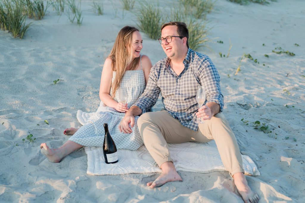 Engaged couple sit on blanket on sand and have champagne at the Isle of Palms beach