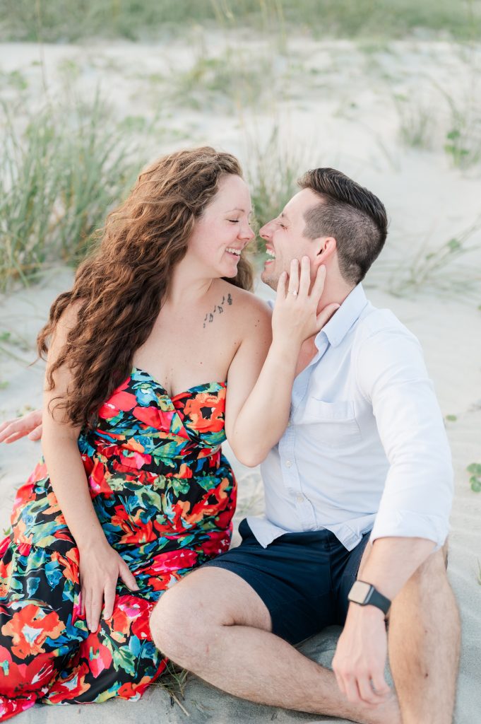 girl holds new fiancé's face in hand as they sit in the sand