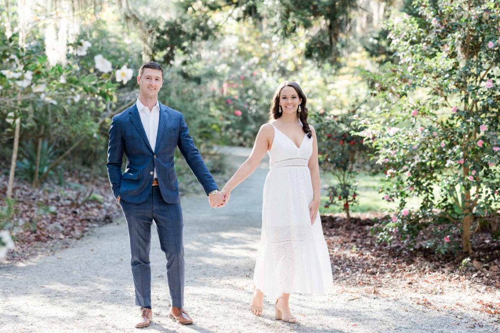 couple holds hands on pathway and look directly into camera