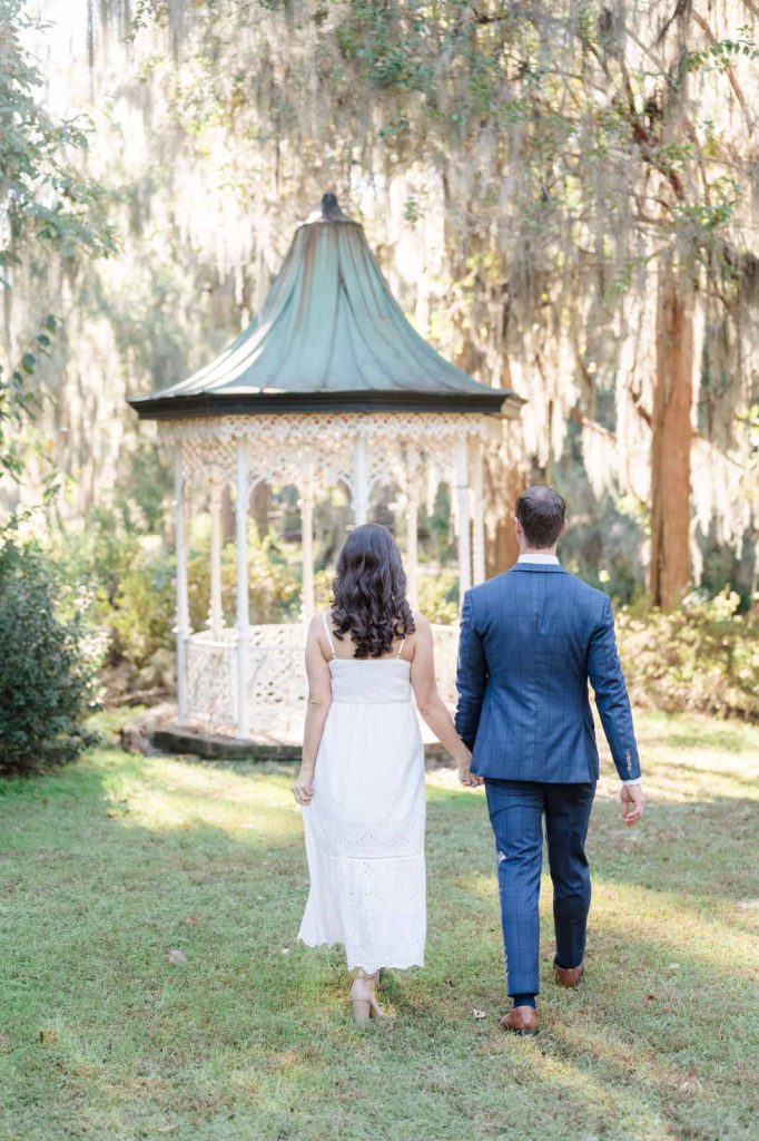 engaged couple walk hand in hand toward white iron gazebo