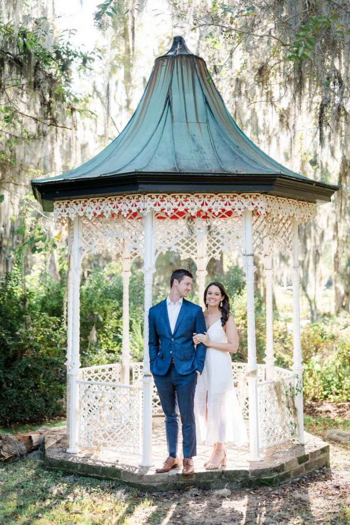 engaged couple stand inside of white iron gazebo in garden