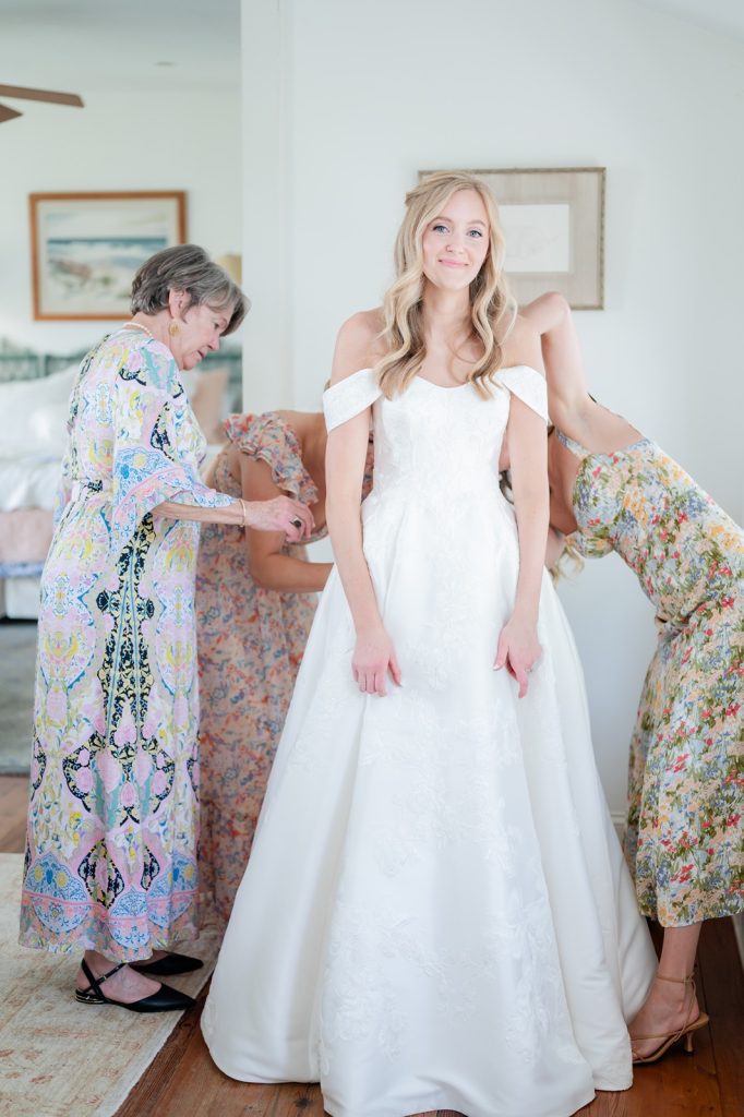bride stands as bridesmaids and mother help zip her into her dress