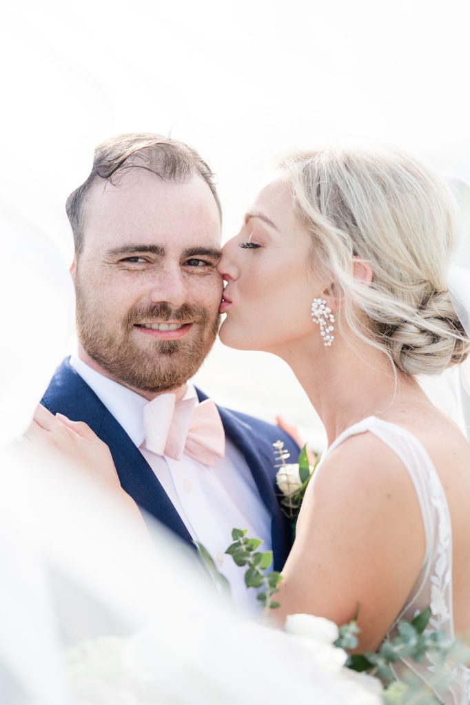 bride kissing cheek of groom while he looks straight into camera