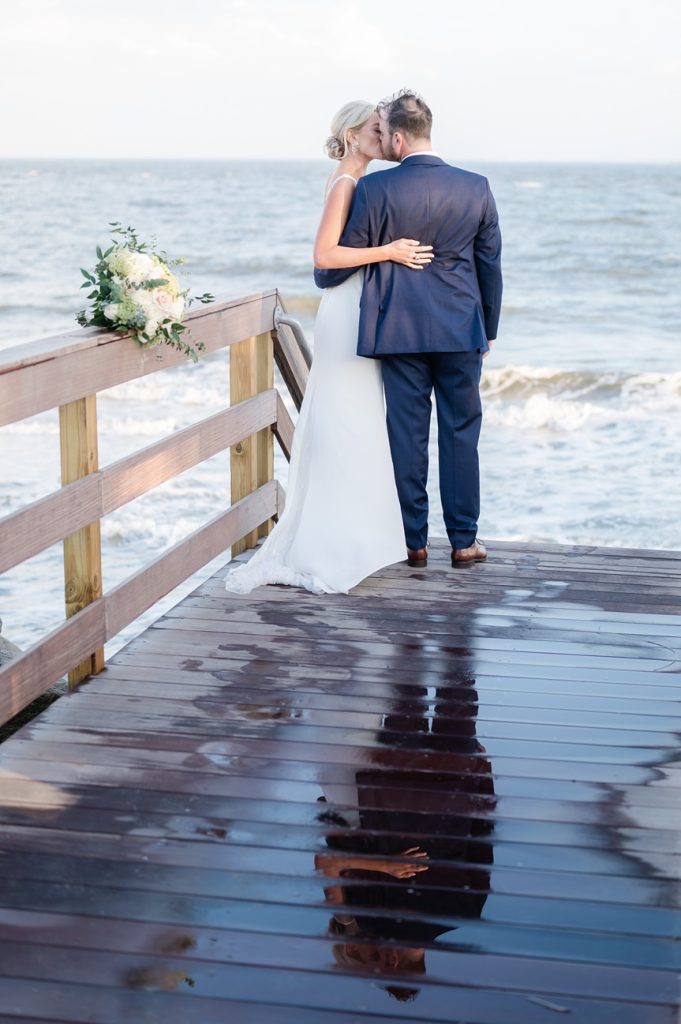 newlywed couple kiss on wet boardwalk overlooking beach, their refection can be seen in boardwalk