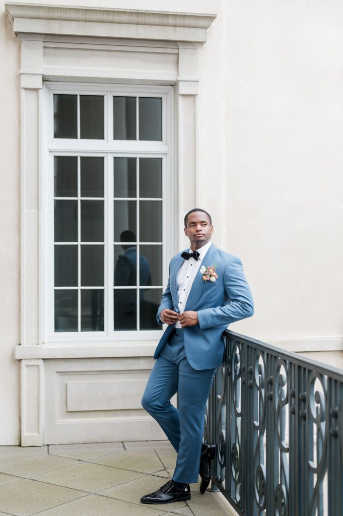 groom in blue tuxedo leaning on iron railing on balcony