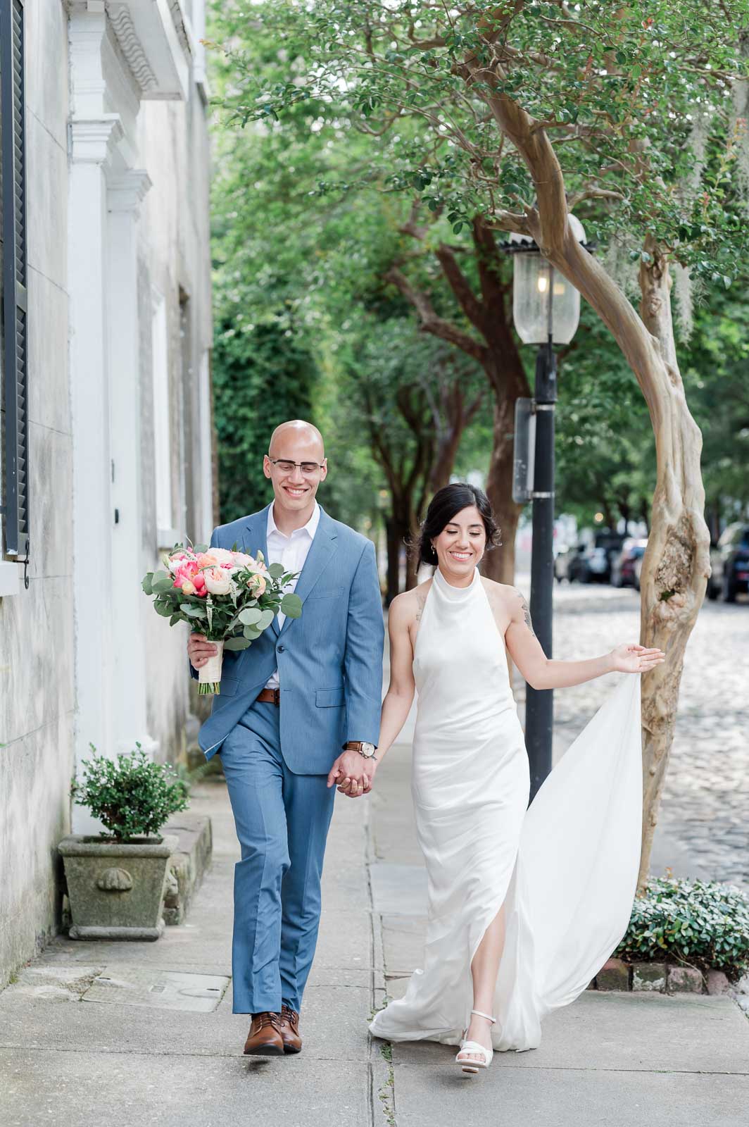 newlyweds walk down street hand in hand with bride holding dress and groom holding flowers