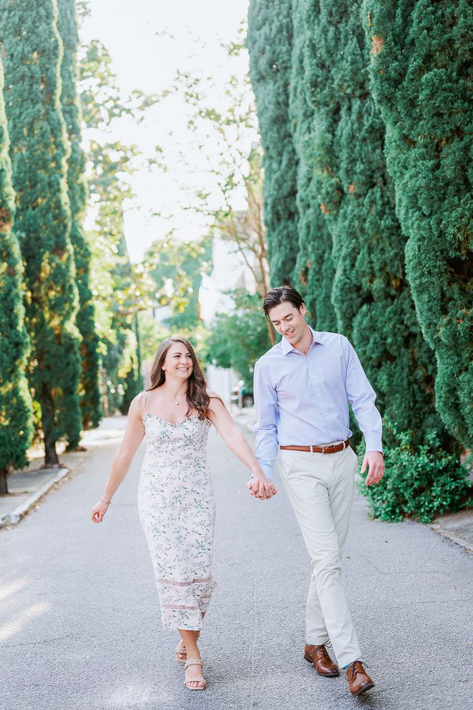 couple hold hands walking down street with cypress trees in the background