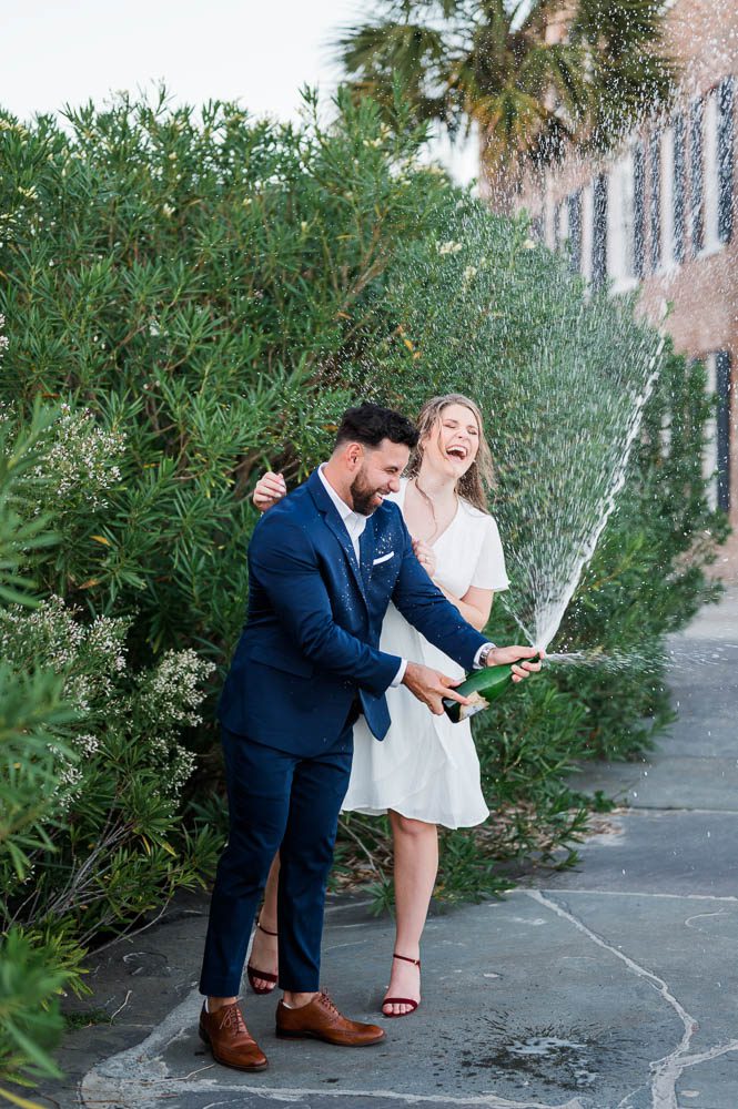 Newly engaged couple pop bottle of champagne in front of bushes