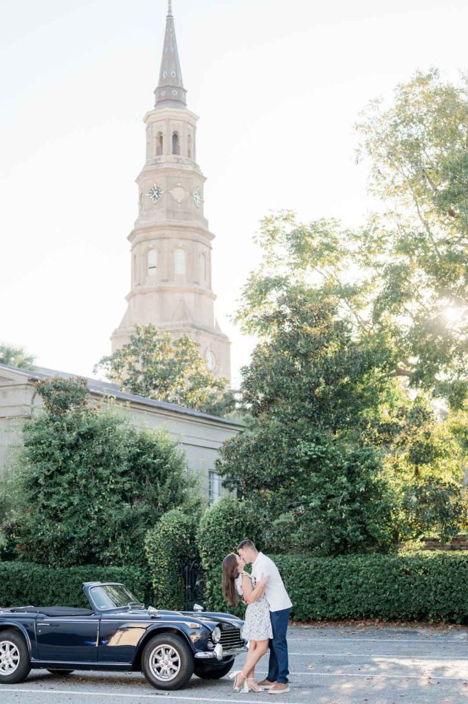 engaged couple kiss in front of blue vintage car and church steeple