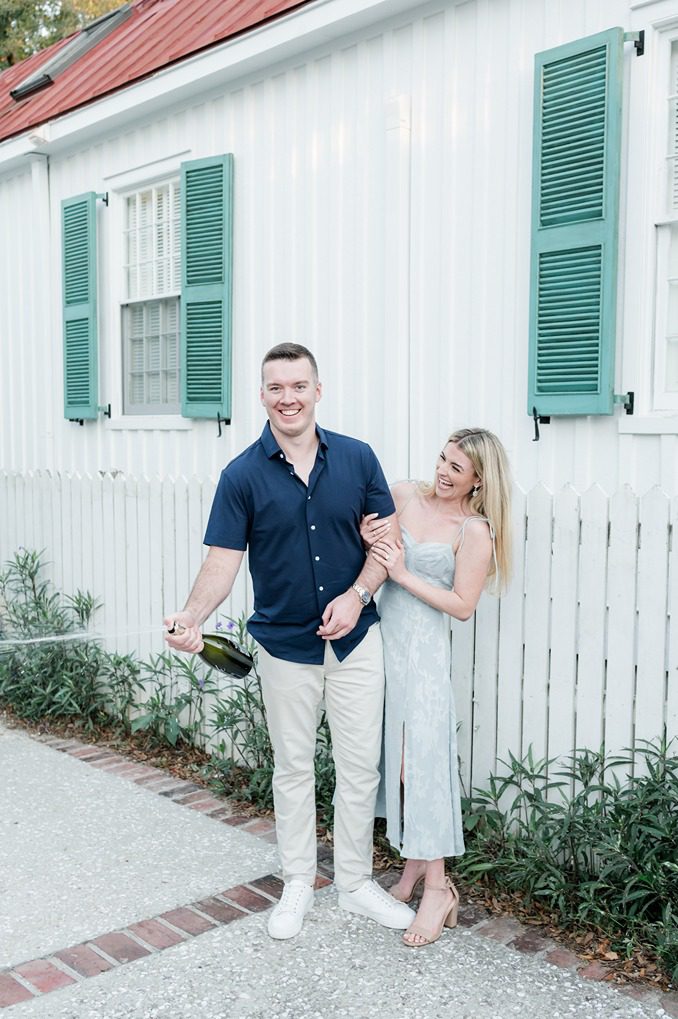 guy stands and sprays bottle of champagne while girlfriend holds his arm and laughs