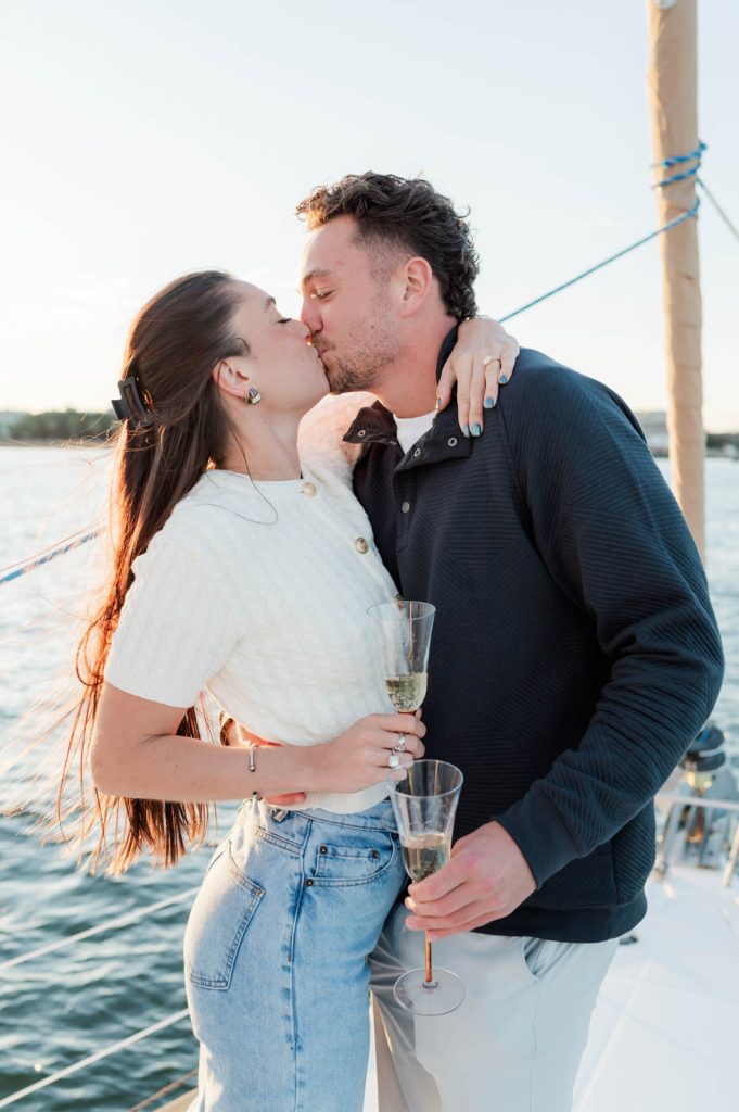Couple kiss after getting engaged on a sailboat, while holding flutes of champagne