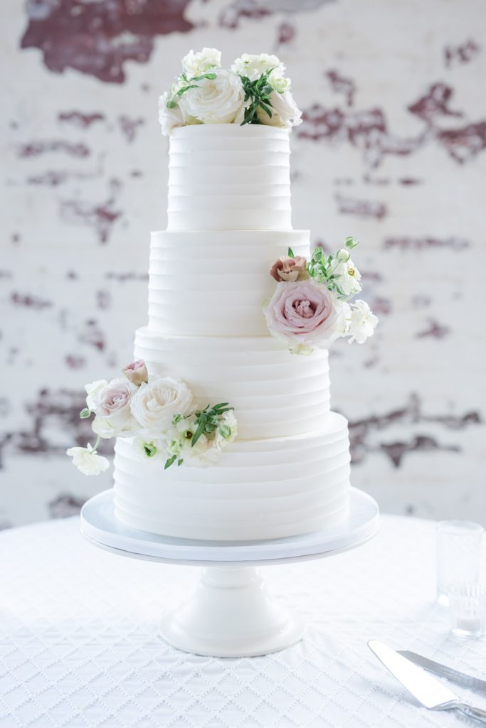 white four layered wedding cake decorated with roses on top of white display plate and white tablecloth