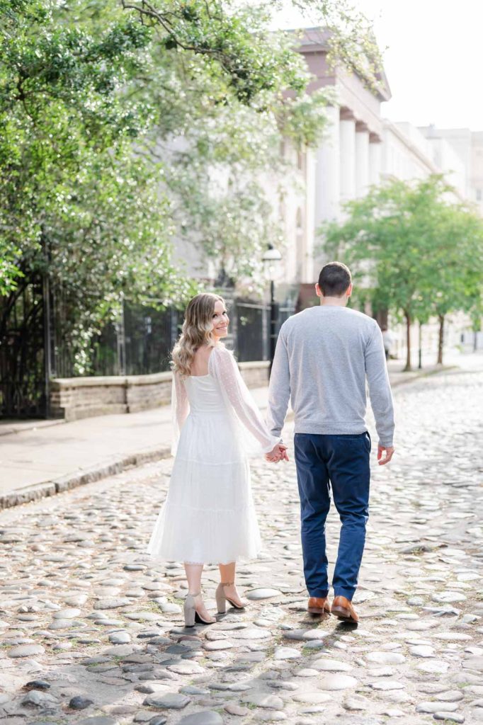 young couple walks away from camera down cobblestone street, girl is looking back at camera over right shoulder