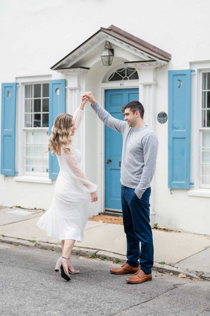 engaged couple twirl in front of white house with blue shutters
