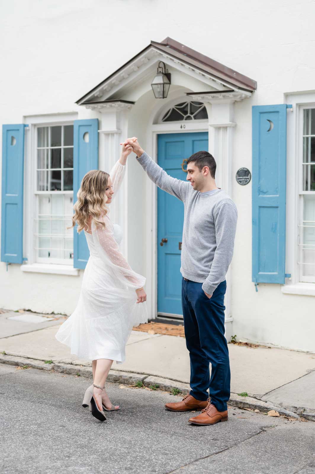 engaged couple twirl in front of white house with blue shutters