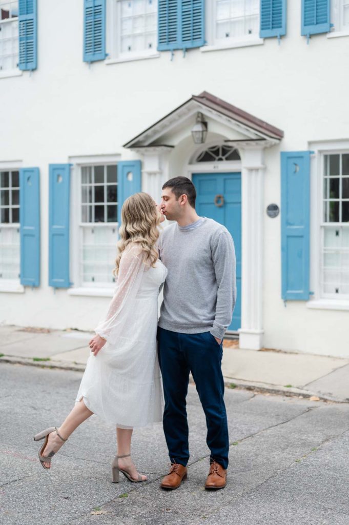 engaged couple kiss in front of white house with blue shutters