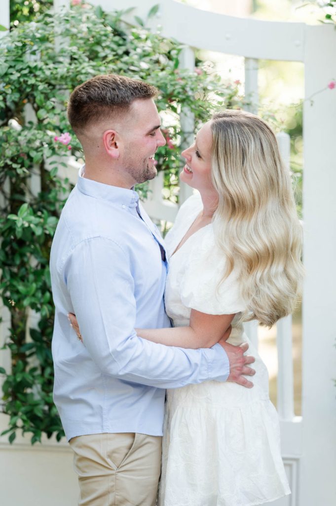engaged couple hold each other around the waist in front of white gate with flowers