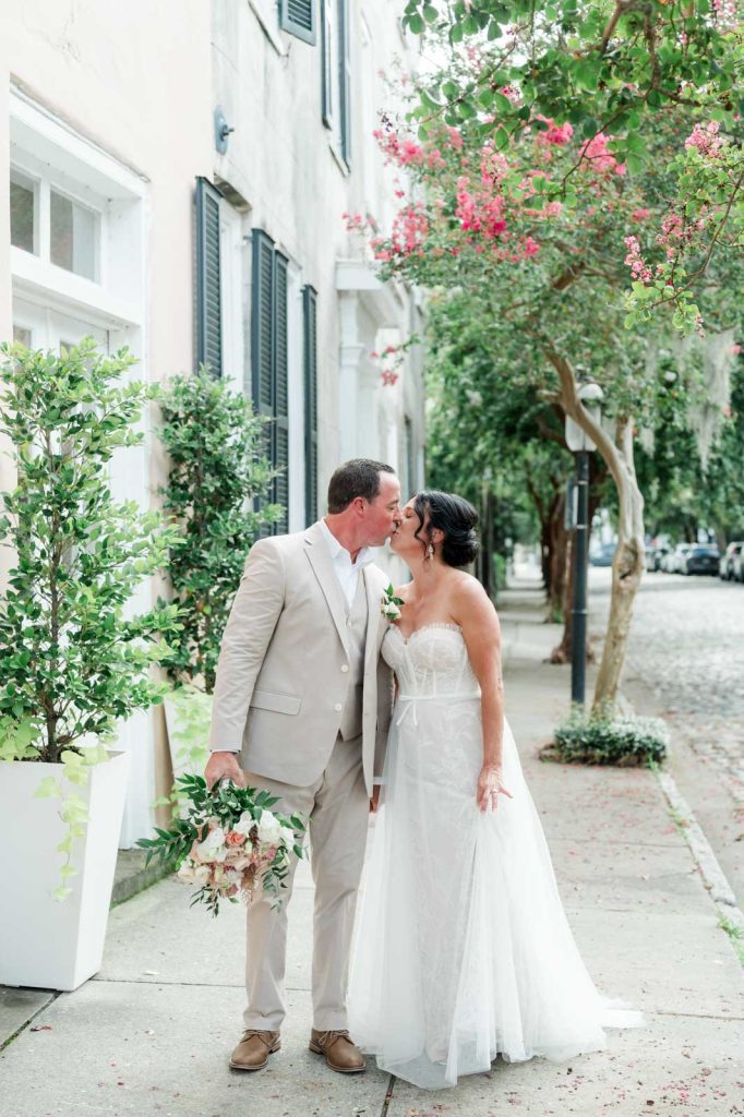 newlyweds kiss on sidewalk with trees in bloom with pink flowers behind