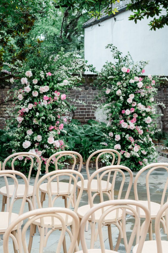 Wedding ceremony setup at Gadsden House featuring a lush floral arch of pink roses and greenery framing wooden chairs arranged for guests in a historic Charleston courtyard setting.