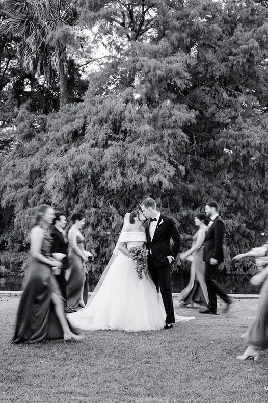 bride and groom kiss in focus while wedding party walks back and forth blurred
