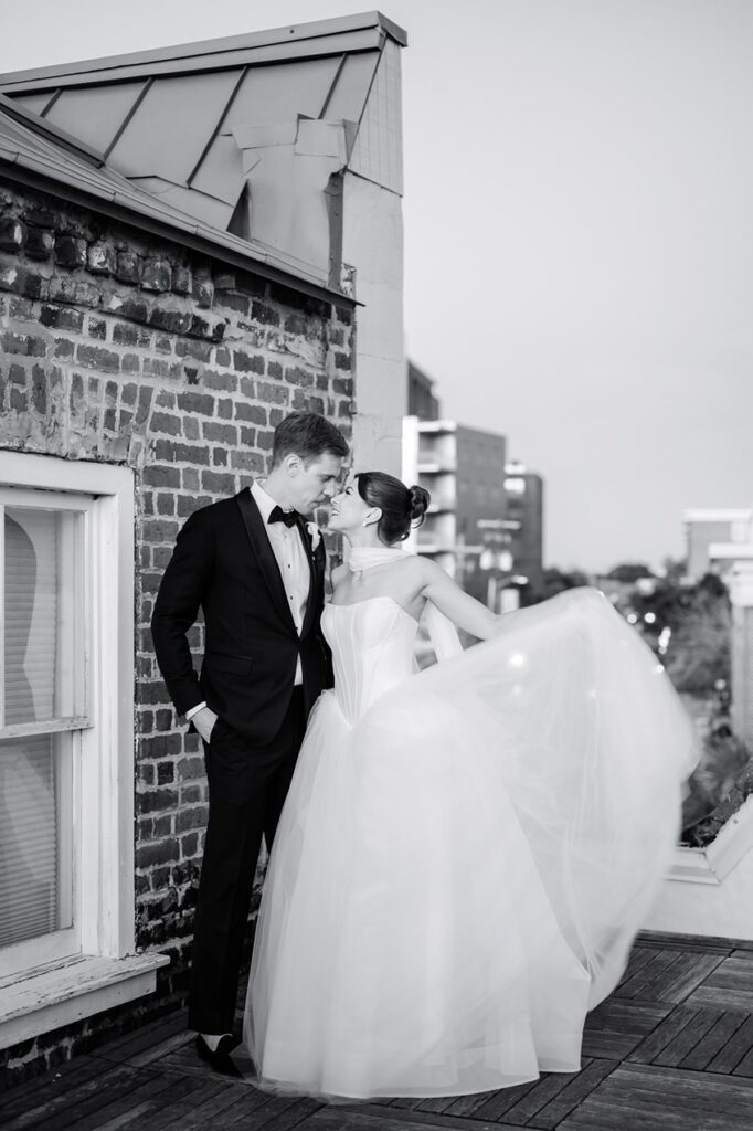 bride and groom stand on rooftop looking into each other's eyes while bride swishes wedding dress