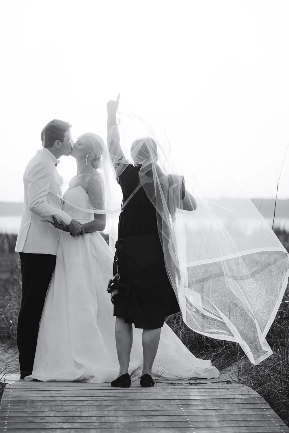 Newlyweds kissing beneath a veil as their photographer Leigh Hayward gently holds it aloft during an intimate wedding moment.