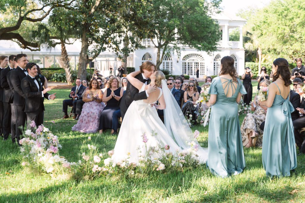 Newlyweds sharing a kiss during their wedding ceremony at Lowndes Grove, surrounded by lush floral arrangements and seated guests in an elegant outdoor setting.