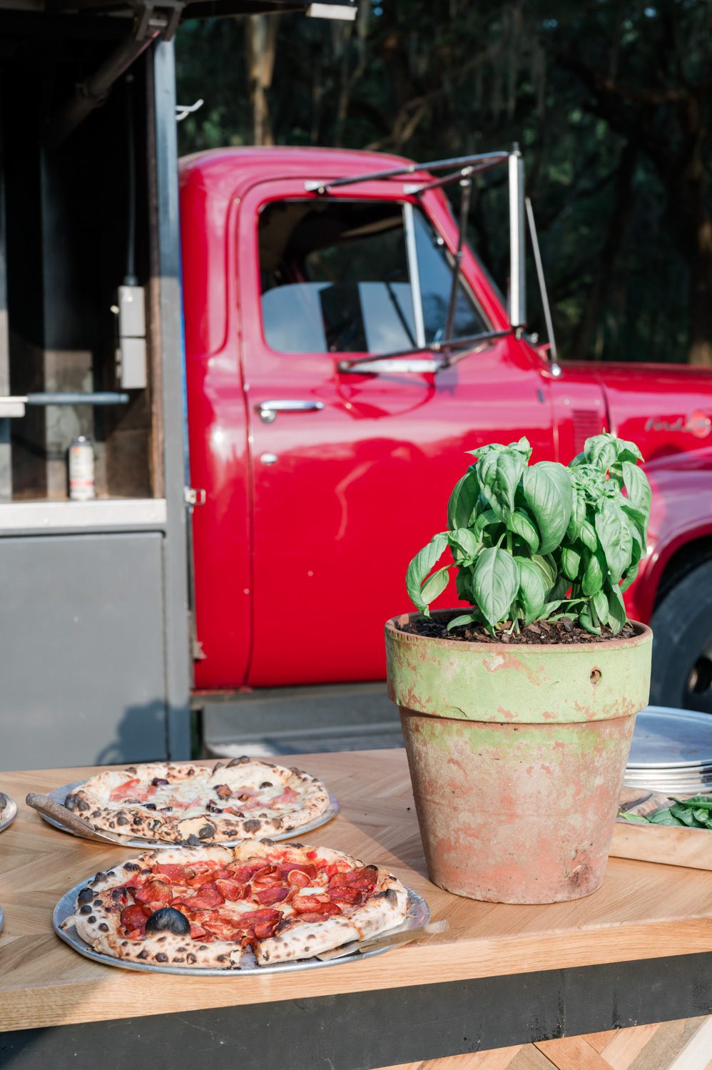Oven-fired pizzas prepared for an Italian-themed rehearsal dinner at Middleton Place, styled with a red food truck in the background and a pot of fresh basil placed beside the pizzas on the serving table.