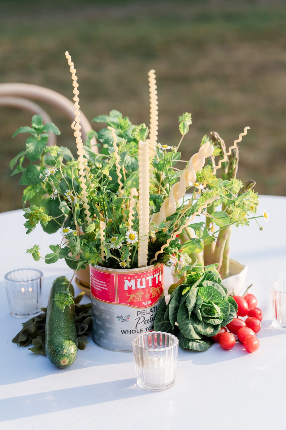 Floral centerpiece arranged in a vintage tomato can, styled with fresh garden vegetables and strands of pasta intermingled throughout the florals, set on an Italian-themed rehearsal dinner table at Middleton Place in Charleston, South Carolina.