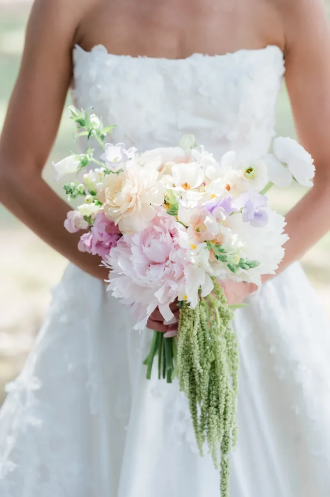 Close-up of a pastel-colored bridal bouquet featuring soft blush, white, lavender, and peach blooms with delicate greenery, held by a bride on her wedding day.