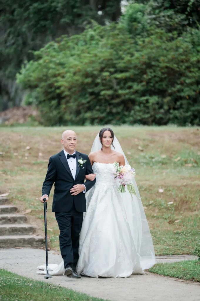 Bride walking arm in arm with her father down the ceremony aisle at Middleton Place, surrounded by the historic garden landscape during a wedding ceremony in Charleston, South Carolina.