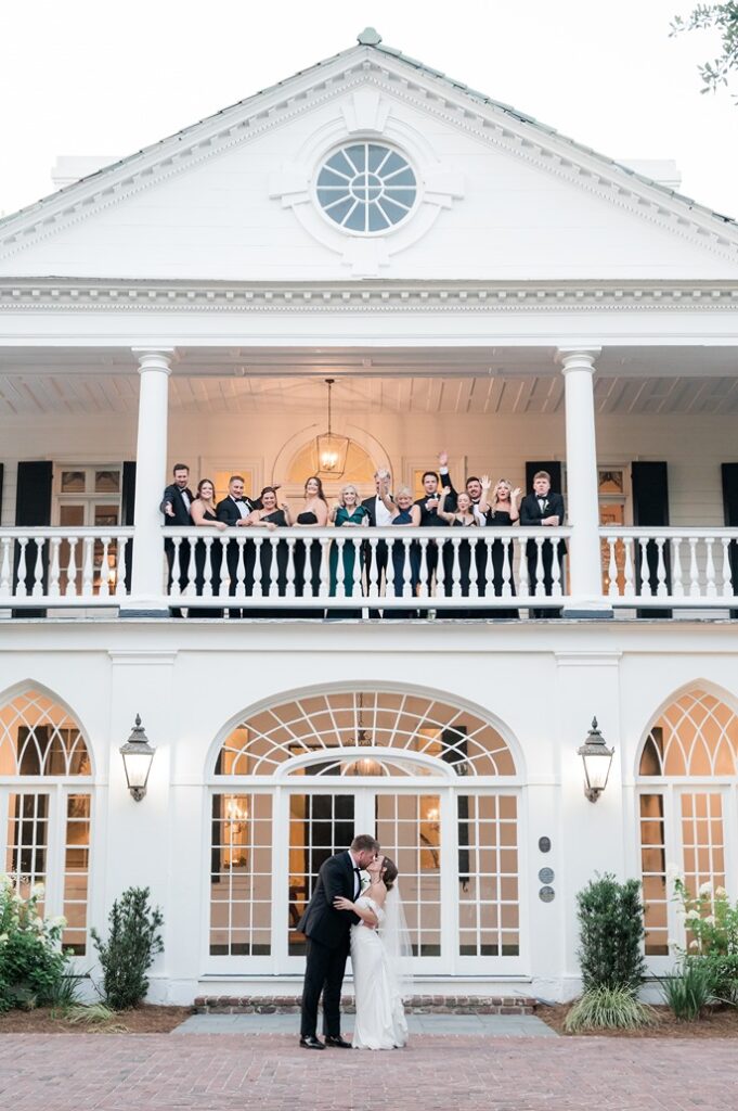 newlyweds kiss on ground level while parents and wedding party cheer from second story porch at Lowndes grove wedding venue